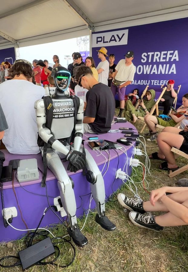 Edek seated among festival-goers at a phone-charging station, plugged into the wall